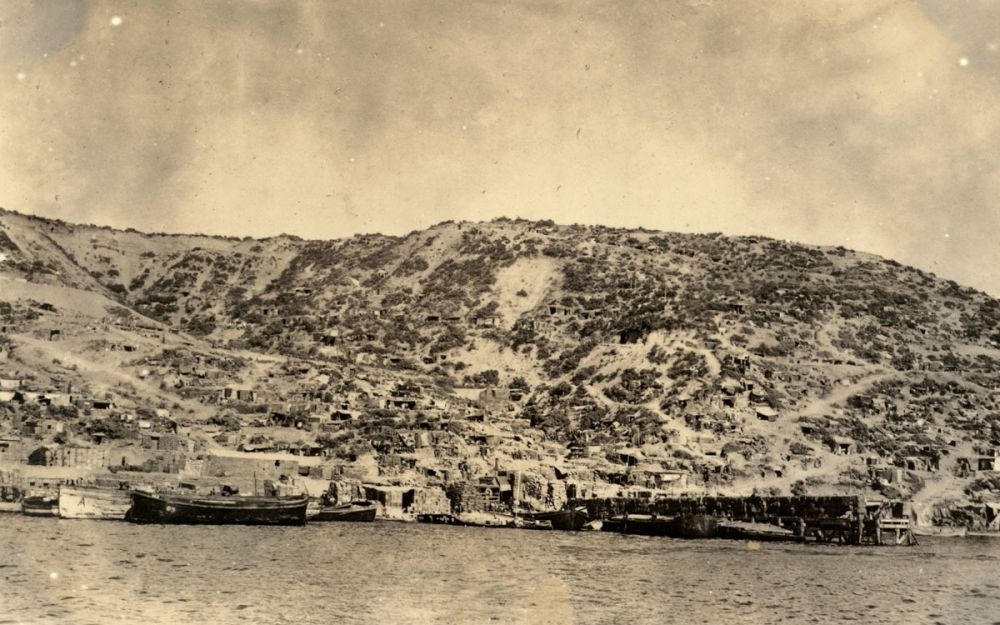 A view of Anzac Cove taken from offshore - a pier, dugouts and stores are visible.